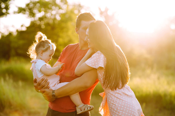 Happy young family walking in the park at sunset. Mom, dad and little daughter having fun in summer park. The concept of a happy family. Parents hold the baby's hands. Kisses and hugs. - Photo, image