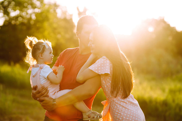 Happy young family walking in the park at sunset. Mom, dad and little daughter having fun in summer park. The concept of a happy family. Parents hold the baby's hands. Kisses and hugs. - Photo, image