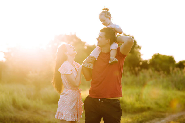 Happy young family walking in the park at sunset. Mom, dad and little daughter having fun in summer park. The concept of a happy family. Parents hold the baby's hands. Kisses and hugs. - Photo, Image
