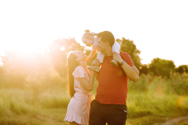 Gelukkige jonge familie wandelen in het park bij zonsondergang. Mam, pap en dochtertje vermaken zich in het zomerpark. Het concept van een gelukkig gezin. Ouders houden de handen van de baby vast. Zoenen en knuffels. - Foto, afbeelding