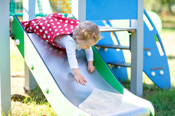 Мила дівчинка, яка грає на слайді на відкритому майданчику. Красива дитина в червоних гумових штанях розважається в сонячний теплий літній день. Дитина ковзає вниз
 - Фото, зображення