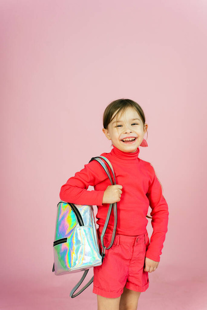 Мила маленька блондинка однокласниця в рожевому костюмі в студії з голографічним рюкзаком. Смішний першокласник на рожевому пастельному фоні з копіспадом
. - Фото, зображення