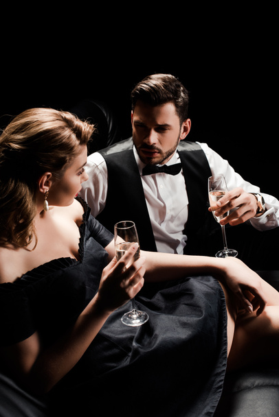 elegante uomo e donna che reggono bicchieri di champagne e si guardano mentre sono seduti isolati sul nero
 - Foto, immagini