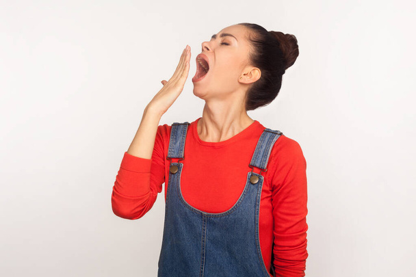 Need to sleep. Portrait of exhausted drowsy girl with hair bun in denim overalls yawning and covering mouth with hand, feeling lazy, suffering insomnia. indoor studio shot isolated on white background - Foto, imagen