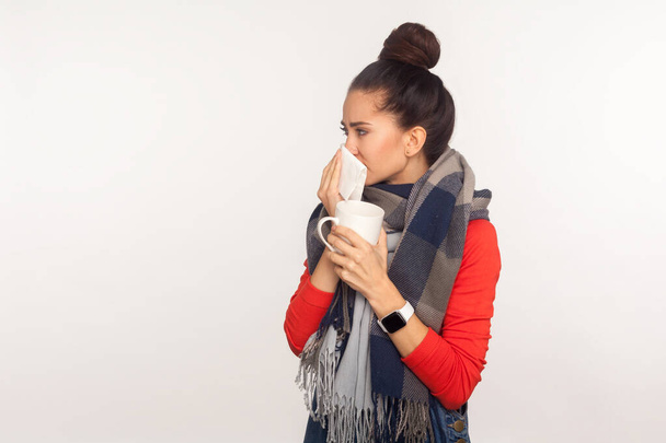 Flu treatment. Portrait of unhealthy young woman wrapped in warm scarf cleaning nose with napkin and holding tea cup, feeling unwell, treating influenza. studio shoot isolated on white background - Foto, immagini