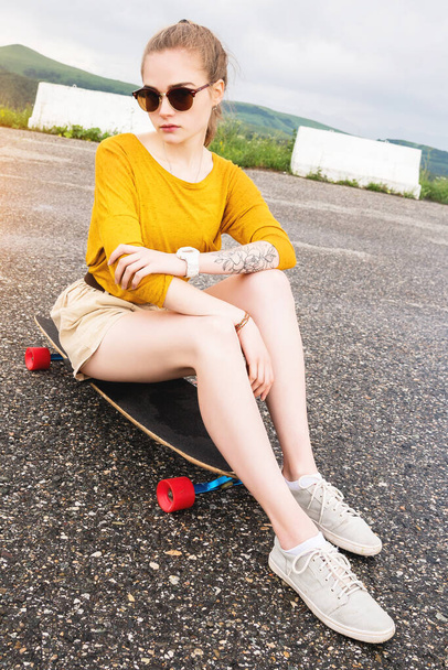 A young attractive girl in a yellow sweater shorts and sunglasses with a tattoo on her arm sits on a longboard behind a suburban asphalt pad against a cloudy sky. - Foto, immagini