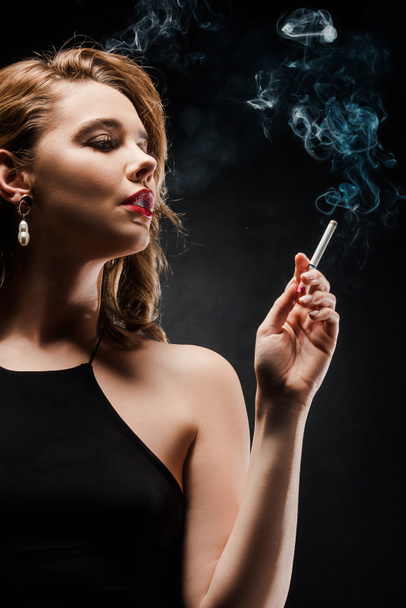 seductive, elegant woman looking away while holding cigarette on black background - Photo, image