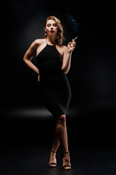full length view of sexy woman in elegant dress holding cigarette while posing on black - Foto, immagini