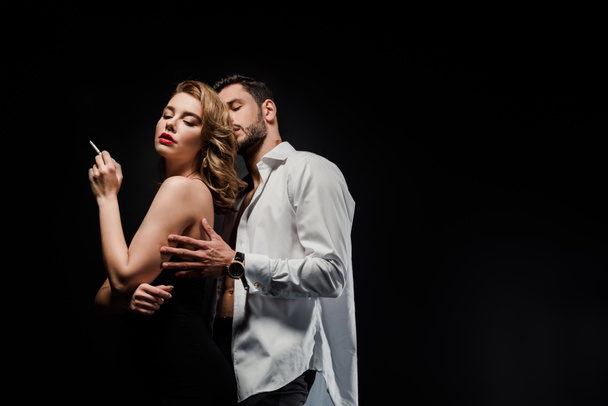 Красивый мужчина в белой рубашке обнимает элегантную, сексуальную женщину, держащую сигарету изолированной на черном
 - Фото, изображение