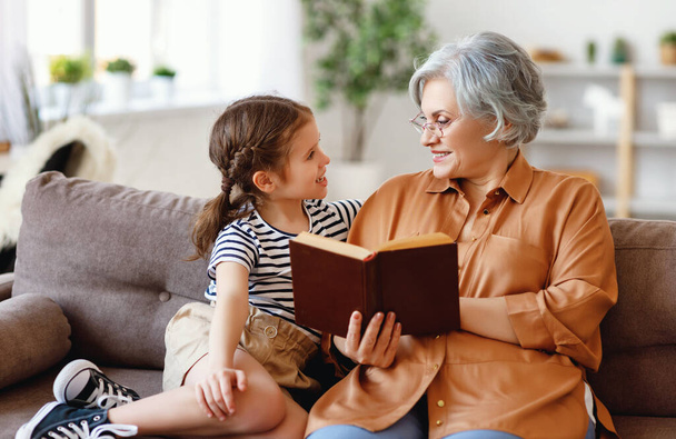 Happy elderly woman and little girl smiling and looking at each other while sitting on couch and reading fascinating fairytale together at hom - Foto, Bild