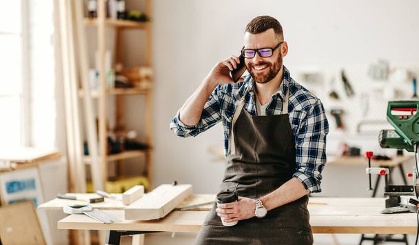 happy young man in apron with cup of takeaway coffee in hand talking on smartphone while leaning on workbench in modern worksho - Photo, Image