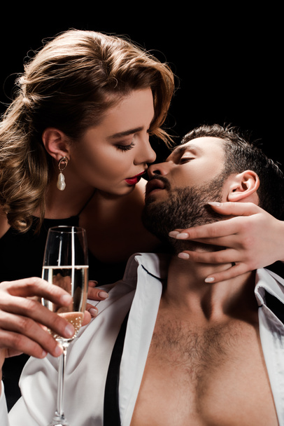 attractive, elegant woman holding champagne glass and kissing handsome man isolated on black - Foto, Imagem