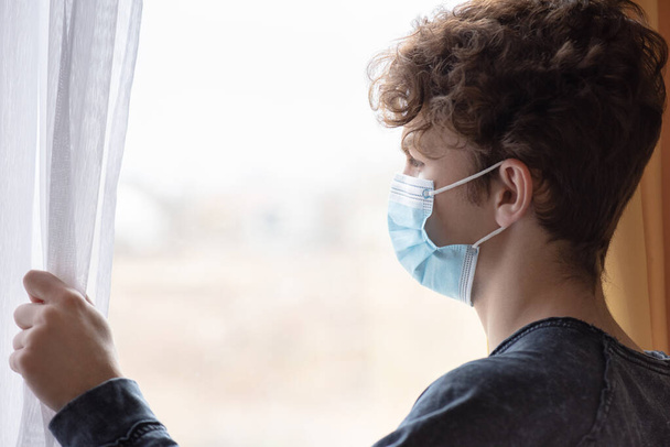 Concept of coronavirus quarantine. Child wearing medical protective face mask during flu virus, looking out of window. COVID-19 - self isolation. Teen boy forced to stay at home. - Foto, imagen
