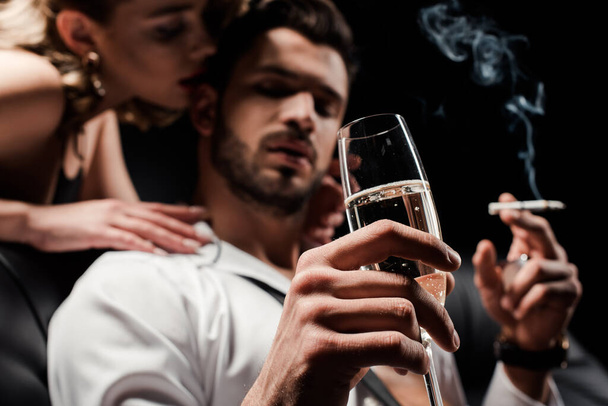selective focus of seductive woman touching handsome, confident man holding champagne glass and smoking isolated on black - Foto, afbeelding