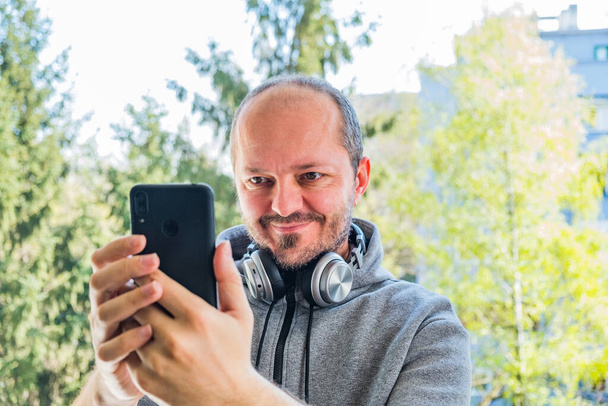 Γενειοφόρος ενήλικας με κουκούλα με ασύρματα ακουστικά που βγάζει selfie με έξυπνο τηλέφωνο σε εξωτερικό χώρο στάθμευσης, κεφάλι και ώμους - Φωτογραφία, εικόνα