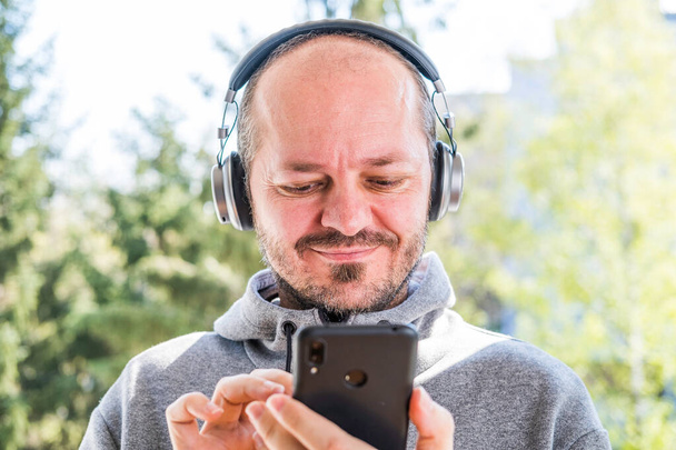 Γενειοφόρος ενήλικας με κουκούλα με ασύρματα ακουστικά που βγάζει selfie με έξυπνο τηλέφωνο σε εξωτερικό χώρο στάθμευσης, κεφάλι και ώμους - Φωτογραφία, εικόνα