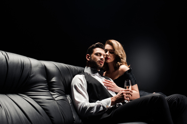 attractive, sexy woman hugging handsome, elegant man sitting on leather sofa with champagne glass on black - 写真・画像