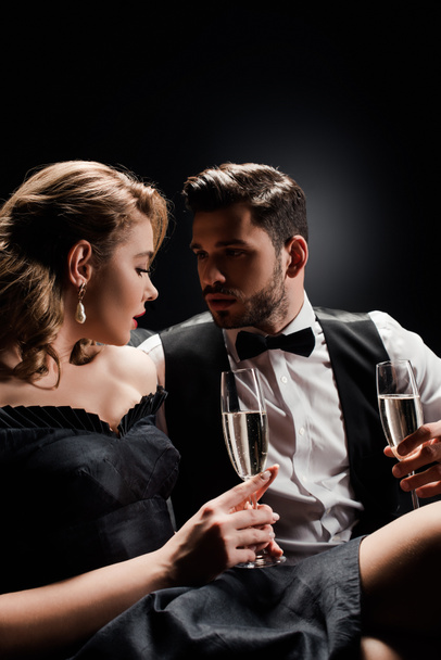 sexy woman and elegant man holding glasses of champagne and looking at each other on black background - Photo, Image