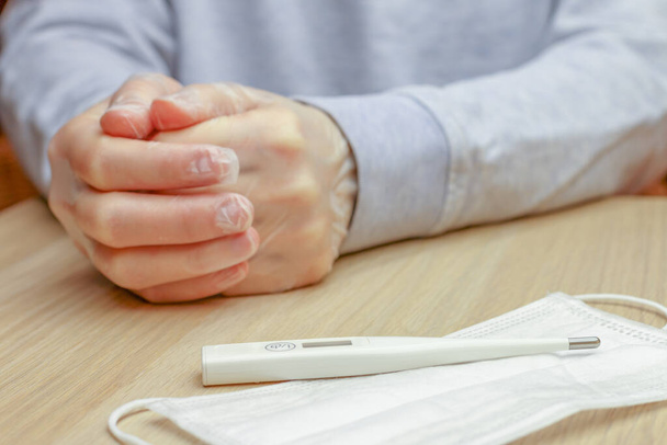 a white disposable protective face mask and a white digital thermometer lying on a table in front of a person sitting with hands in clean protective gloves, idea of protection and self care - Photo, Image