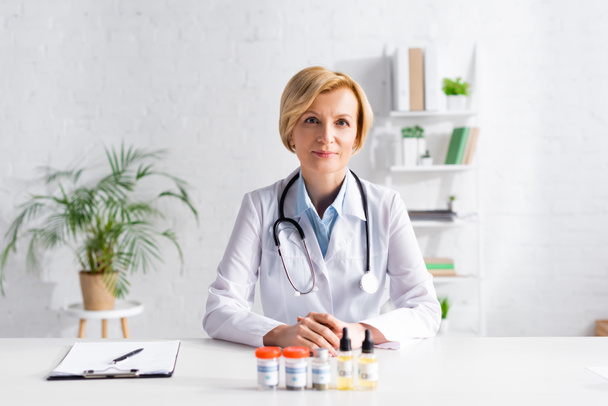 mature doctor sitting at desk with bottles of cbd near clipboard  - Photo, Image