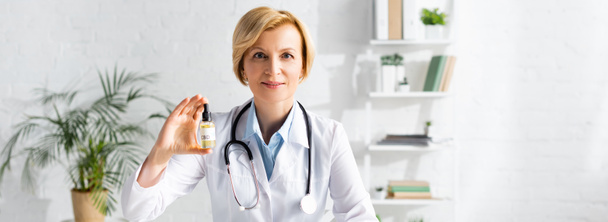 Panoramaaufnahme eines reifen Arztes in weißem Mantel, der eine Flasche mit cbd-Schriftzug in der Klinik hält  - Foto, Bild