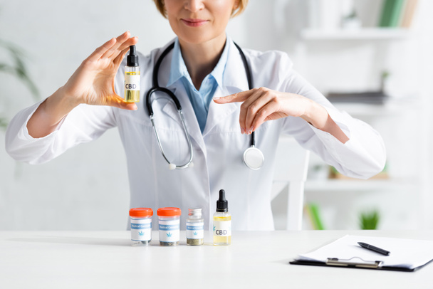 cropped view of doctor in white coat pointing with finger at bottle with cbd lettering  - Fotografie, Obrázek