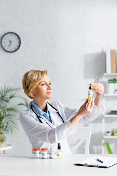 atrakcyjny i dojrzały lekarz w białym płaszczu trzymający butelkę z napisem cbd w klinice  - Zdjęcie, obraz
