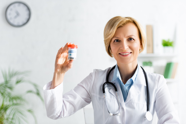 happy and mature doctor in white coat holding bottle with medical cannabis lettering  - Foto, afbeelding