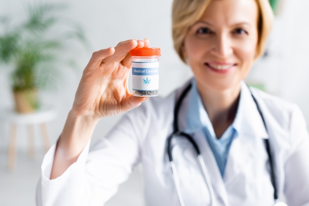 selective focus of cheerful and mature doctor in white coat holding bottle with medical cannabis lettering  - Photo, Image