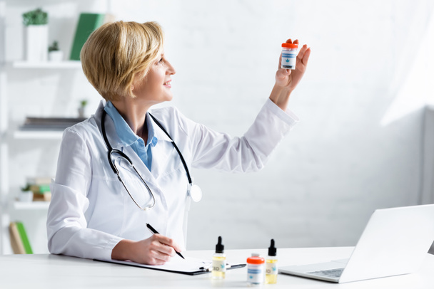 happy and mature doctor in white coat looking at bottle with medical cannabis lettering  - Fotografie, Obrázek