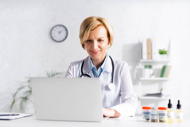 medico felice utilizzando laptop vicino a bottiglie con cannabis medica e cbd
  - Foto, immagini