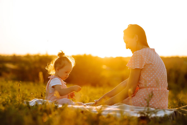 Mother and little daughter playing together in a summer park at sunset. Happy mom with small child  having fun on sunny field. Kisses and hugs. The concept of a happy family.  - Фото, зображення