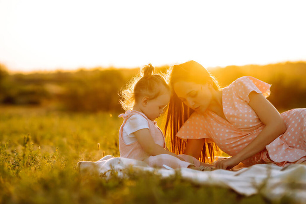 Anne ve küçük kızı gün batımında bir yaz parkında birlikte oynuyorlar. Küçük çocuklu mutlu bir anne güneşli bir alanda eğleniyor. Öpücükler ve sarılmalar. Mutlu aile kavramı..  - Fotoğraf, Görsel