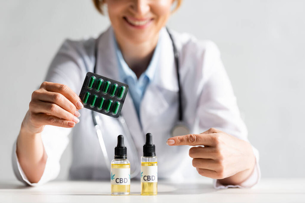 cropped view of happy and mature doctor in white coat pointing with finger at bottle with cbd lettering and holding blister pack with pills in clinic  - Foto, Imagem