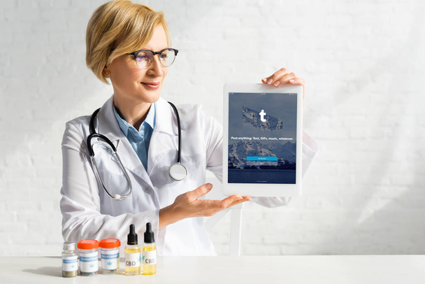 KYIV, UKRAINE - MARCH 5, 2020: mature doctor in white coat holding digital tablet with tumblr app near bottles with cbd and medical cannabis lettering - Foto, Imagen