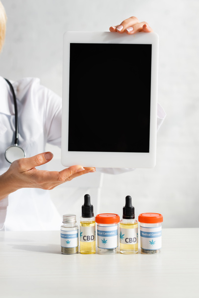 cropped view of doctor holding digital tablet with blank screen near bottles with cbd and medical cannabis lettering in clinic  - Foto, afbeelding