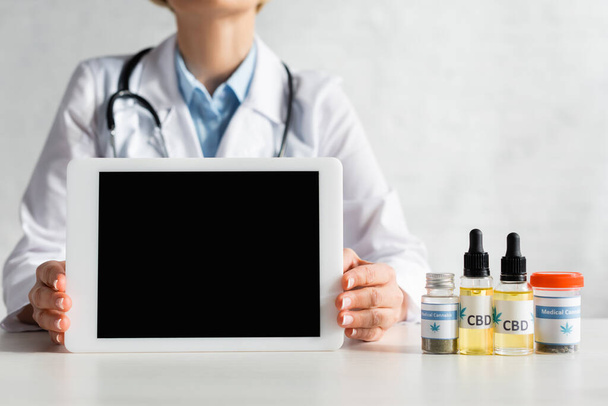 cropped view of mature doctor in white coat holding digital tablet with blank screen near bottles with cbd and medical cannabis lettering in clinic  - Foto, Imagen