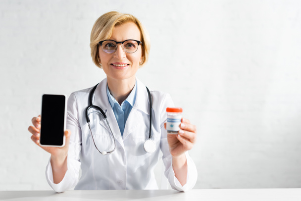 happy and mature doctor in white coat holding smartphone with blank screen and bottle with medical cannabis lettering  - Photo, Image