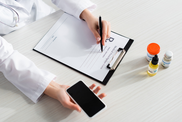 обрезанный вид врача, выписывающего рецепт рядом с бутылками с CBD и медицинской марихуаной, держа смартфон с пустым экраном
  - Фото, изображение