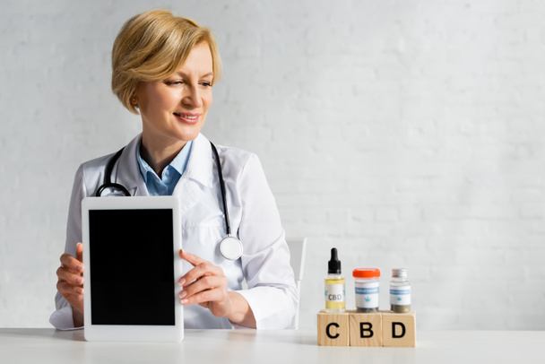 щасливий лікар тримає цифровий планшет з порожнім екраном біля кубиків з пляшками та наркотиками
  - Фото, зображення