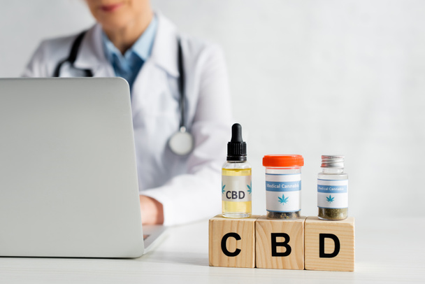 cropped view of doctor using laptop near cubes with cbd lettering and bottles with medical cannabis  - Foto, Imagem