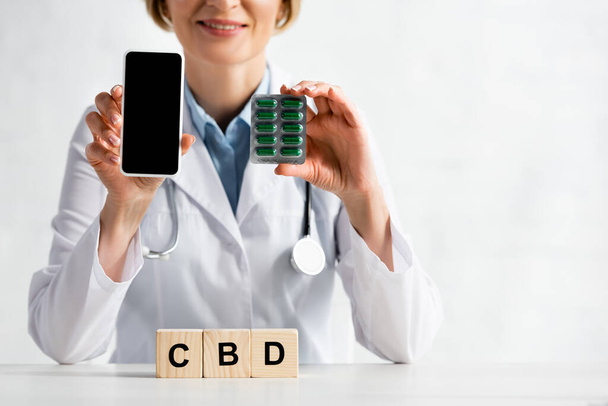 cropped view of happy doctor holding smartphone with blank screen and blister pack with pills near cubes with cbd lettering  - Photo, image