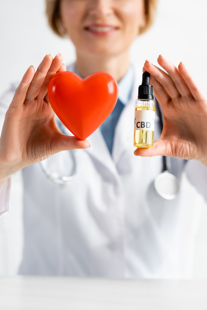cropped view of happy and mature doctor holding bottle with cbd lettering and red heart in clinic  - 写真・画像
