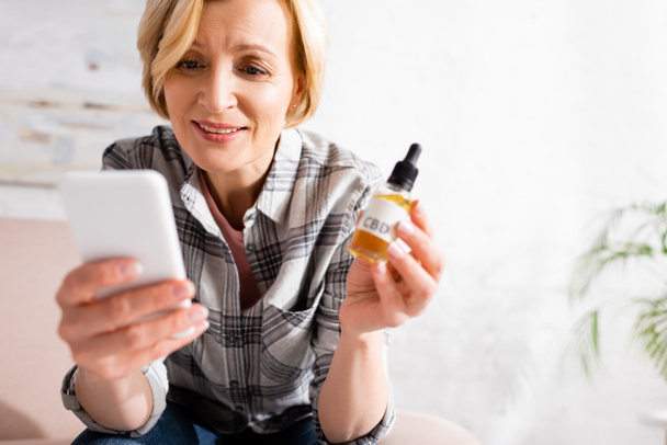 selective focus of happy and mature woman holding bottle with cbd lettering and using smartphone  - Photo, image