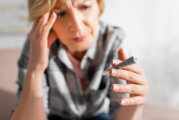 enfoque selectivo de la mujer madura con dolor de cabeza mirando conjunta con marihuana legal en el hogar
  - Foto, Imagen