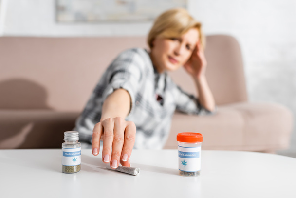 selective focus mature woman taking rolled joint with legal marijuana near bottles with medical cannabis lettering - Φωτογραφία, εικόνα