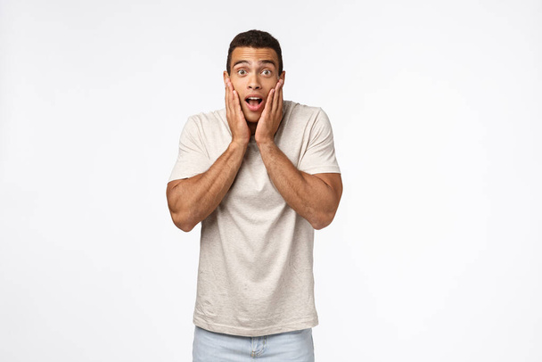 Shocked, astonished young muscline man look speechless and fascinated, grab face from amazement, drop jaw and stare camera excited, see something brand new and cool, standing white background - Photo, Image