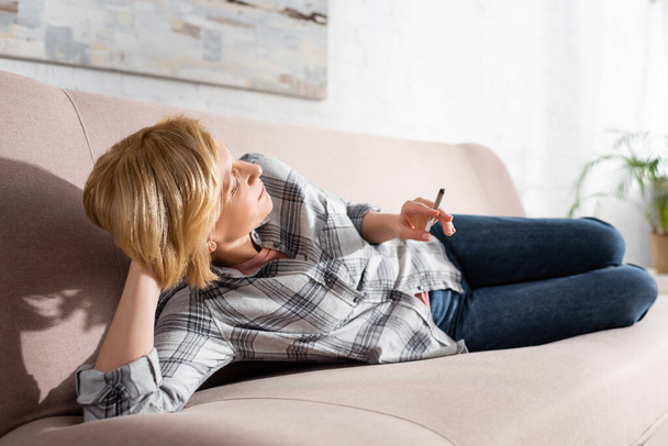 mature woman lying on sofa and holding joint with legal marijuana  - 写真・画像