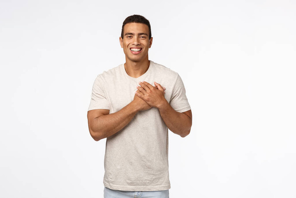 Guy feeling touched and flattered receiving praises. Charming hispanic young man in t-shirt, press hands to heart, smiling and laughing carefree, express gratitude, white background - 写真・画像