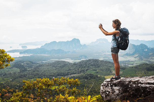 Young lady hiker with backpack standing on top of the mountain and taking a picture of valley - Foto, Imagem